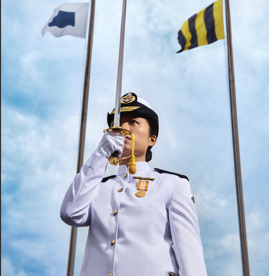 Naval officer with binoculars on bridge, with two other officers at controls in the background.